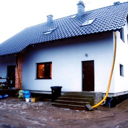 Mahon - Inexo - Nowo otynkowany dom jednorodzinny z ciemną dachówką, widoczne okno z ciepłym światłem wewnątrz, brązowe drzwi wejściowe i żółty wąż prawdopodobnie do odprowadzania wody po myciu elewacji.