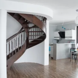 Wnętrze domu z krętymi, drewnianymi schodami z metalowymi poręczami, widoczne drzwi z przeszkleniami, kuchnia z białymi szafkami i pomarańczowy fotel.