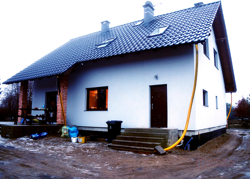 Nowo otynkowany dom jednorodzinny z ciemną dachówką, widoczne okno z ciepłym światłem wewnątrz, brązowe drzwi wejściowe i żółty wąż prawdopodobnie do odprowadzania wody po myciu elewacji.