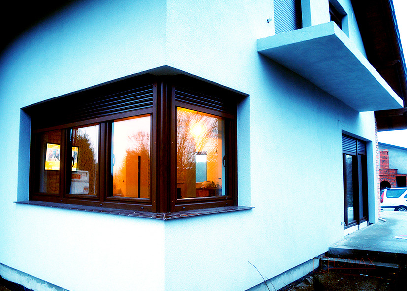 Narożne okno z brązową ramą na elewacji domu, z widocznym odbiciem drzew i wnętrza w szybach, z żaluzjami zewnętrznymi.