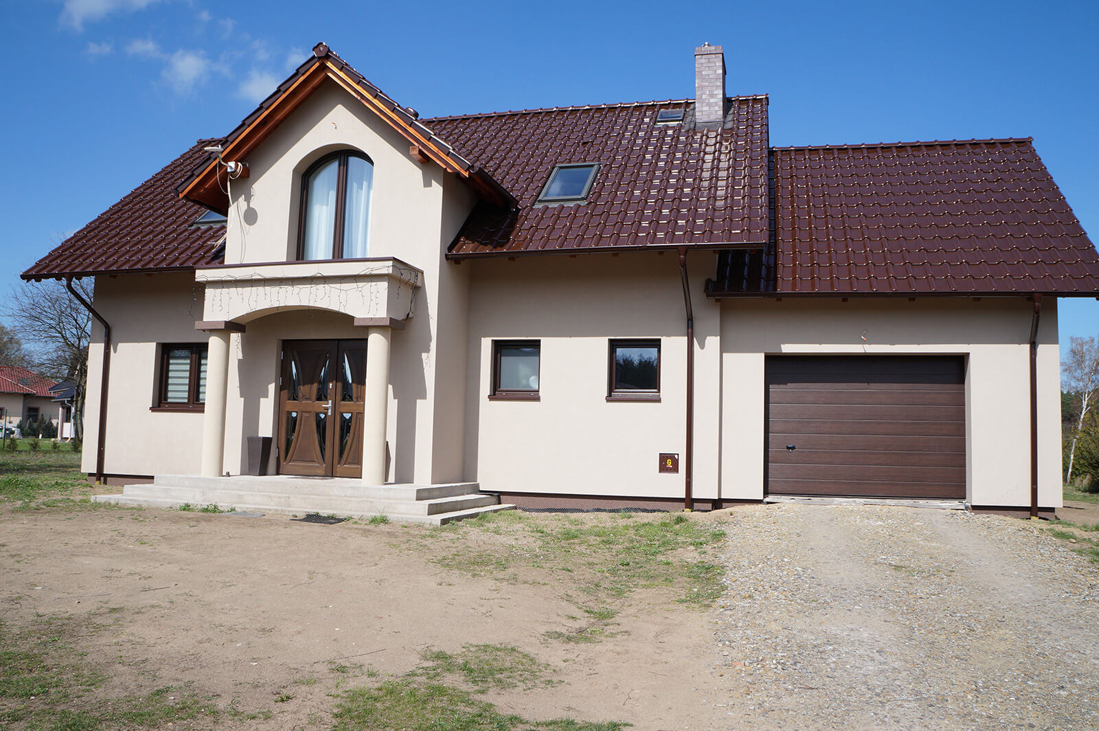 Dom jednorodzinny z brązowym dachem i beżową elewacją, widoczne okna z brązowymi ramami, dwuskrzydłowe drzwi wejściowe z kolumnami i brama garażowa segmentowa w kolorze brązowym.