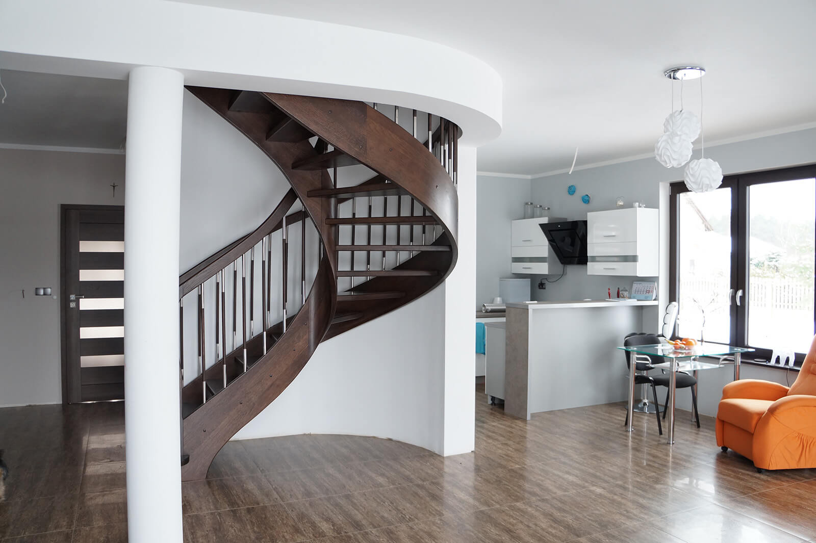 Wnętrze domu z krętymi, drewnianymi schodami z metalowymi poręczami, widoczne drzwi z przeszkleniami, kuchnia z białymi szafkami i pomarańczowy fotel.