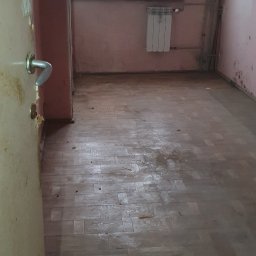 ARSENBUD ANTON SOLOVIOV - Pokój przed cyklinowaniem: zniszczona podłoga parkietowa, ściany z plamami, okno z widokiem na miasto. Widoczne ślady po zalaniach i zabrudzeniach.