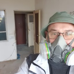 ARSENBUD ANTON SOLOVIOV - Pracownik w masce ochronnej, w trakcie remontu wnętrza z widocznymi drzwiami i oknem. Ujęcie z bliska na twarz, widoczny fragment pomieszczenia w tle.