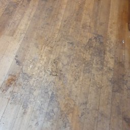 ARSENBUD ANTON SOLOVIOV - Drewniana podłoga przed cyklinowaniem, widoczne zabrudzenia i zarysowania. Fragment nogi od krzesła i buta. Podłoga wymaga renowacji.