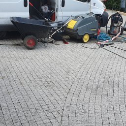 ARSENBUD ANTON SOLOVIOV - Przygotowanie do czyszczenia kostki brukowej: wózek, myjka ciśnieniowa Karcher i rozłożone węże na szarej kostce. W tle biały bus i osoba w kamizelce.