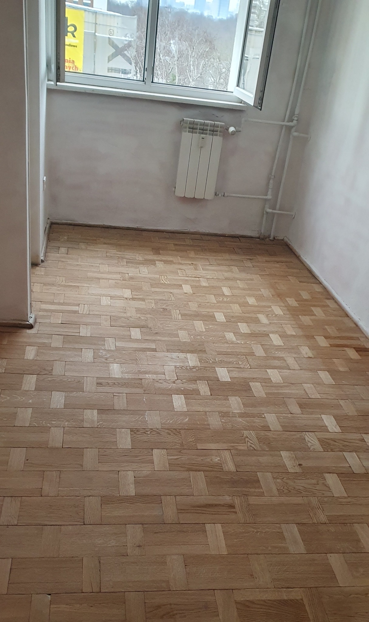 Parkiet ułożony w jodełkę w jasnym pokoju z oknem i grzejnikiem. Świeżo wycyklinowana podłoga z widocznym wzorem drewna.