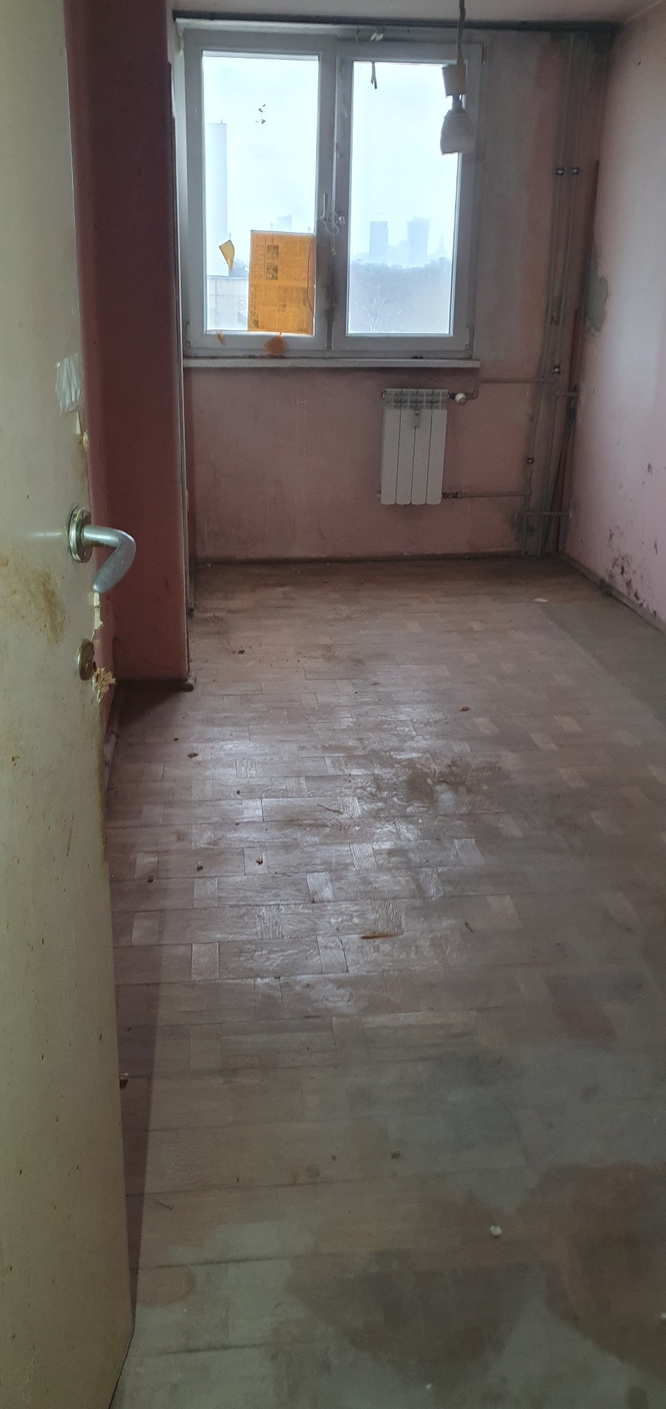 Pokój przed cyklinowaniem: zniszczona podłoga parkietowa, ściany z plamami, okno z widokiem na miasto. Widoczne ślady po zalaniach i zabrudzeniach.