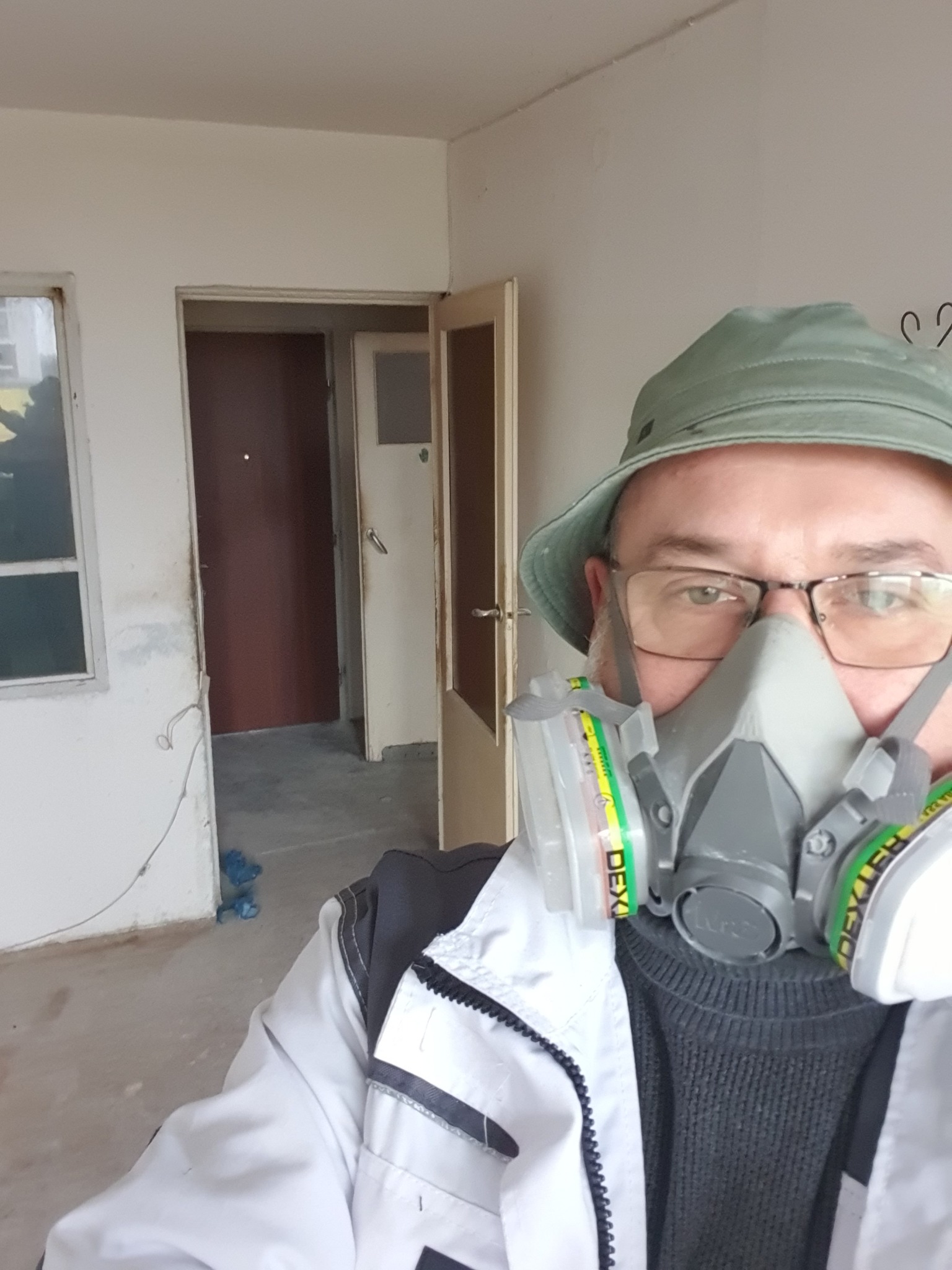 Pracownik w masce ochronnej, w trakcie remontu wnętrza z widocznymi drzwiami i oknem. Ujęcie z bliska na twarz, widoczny fragment pomieszczenia w tle.