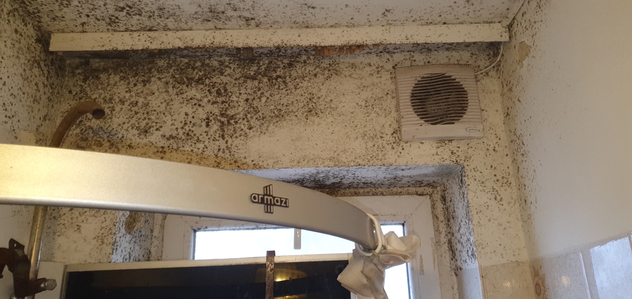 Zaniedbana łazienka z widocznym zagrzybieniem na ścianach, suficie i wokół okna. Brudny wentylator i rura. Widoczna suszarka na pranie.