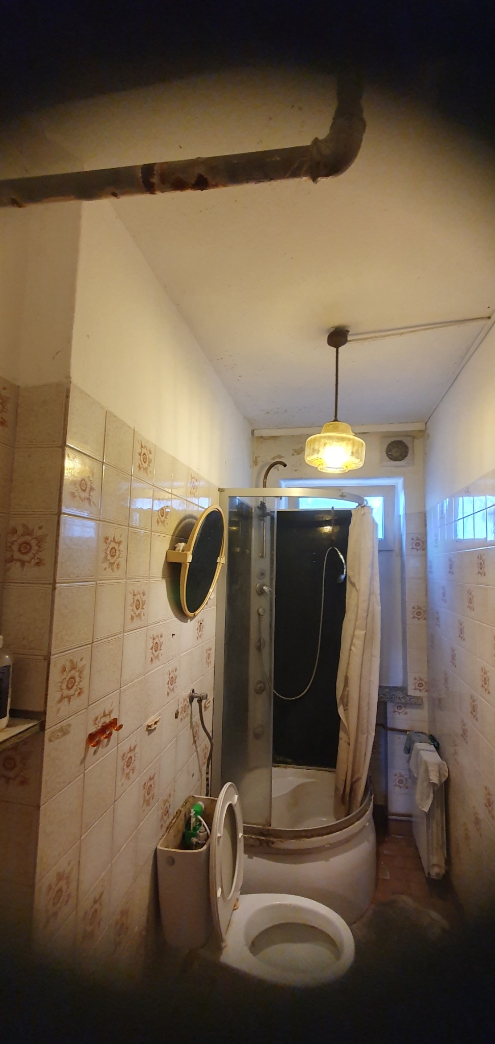 Zaniedbana łazienka z kabiną prysznicową, toaletą z otwartą klapą i rdzewiejącą rurą na suficie. Widoczne ślady pleśni i wilgoci. Potrzebny remont.