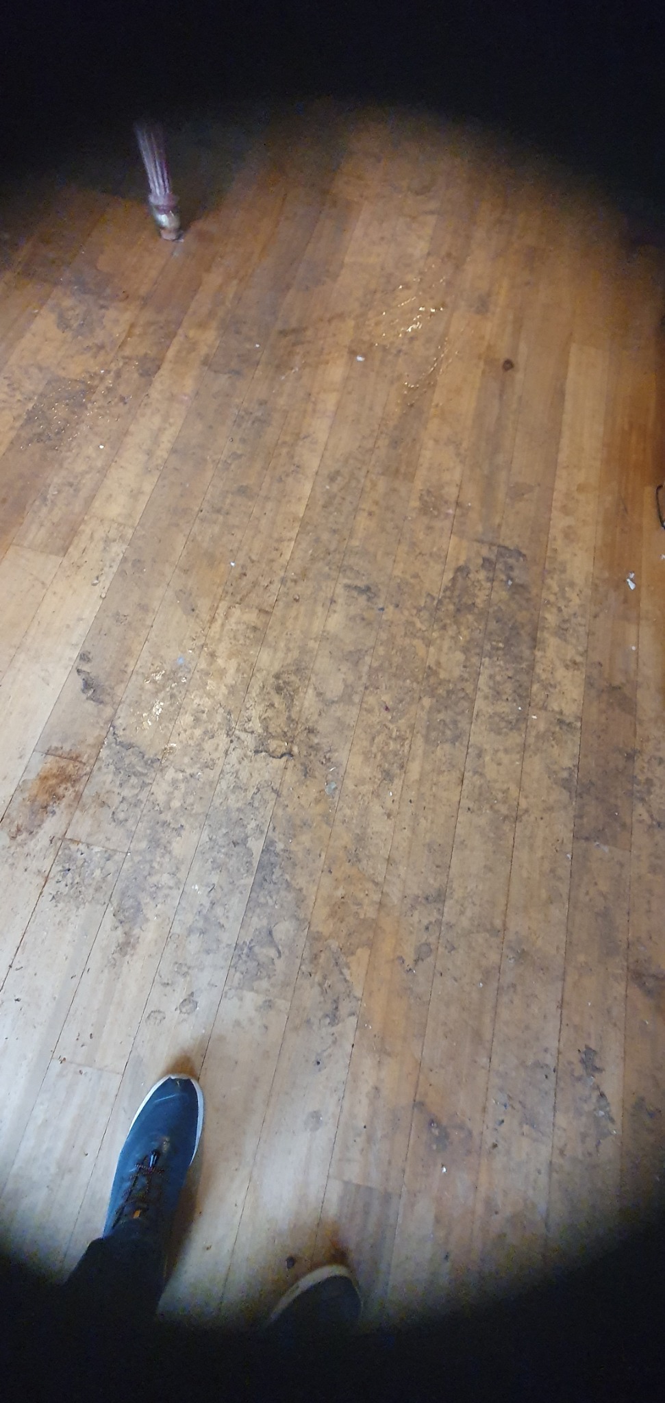 Drewniana podłoga przed cyklinowaniem, widoczne zabrudzenia i zarysowania. Fragment nogi od krzesła i buta. Podłoga wymaga renowacji.