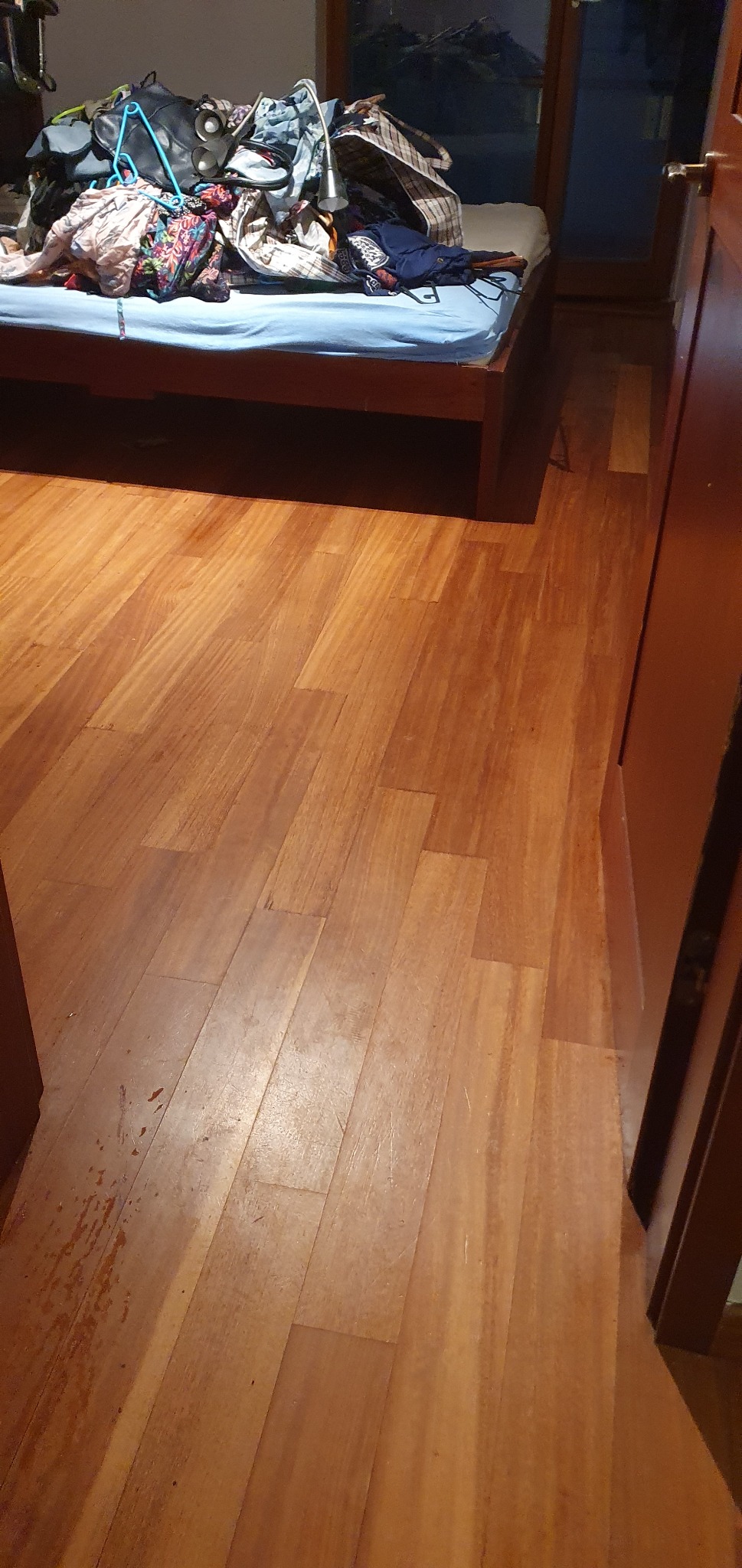 Parkiet z widocznymi rysami i zabrudzeniami. Na łóżku w tle leżą ubrania i torby. Widoczne drzwi i okno.