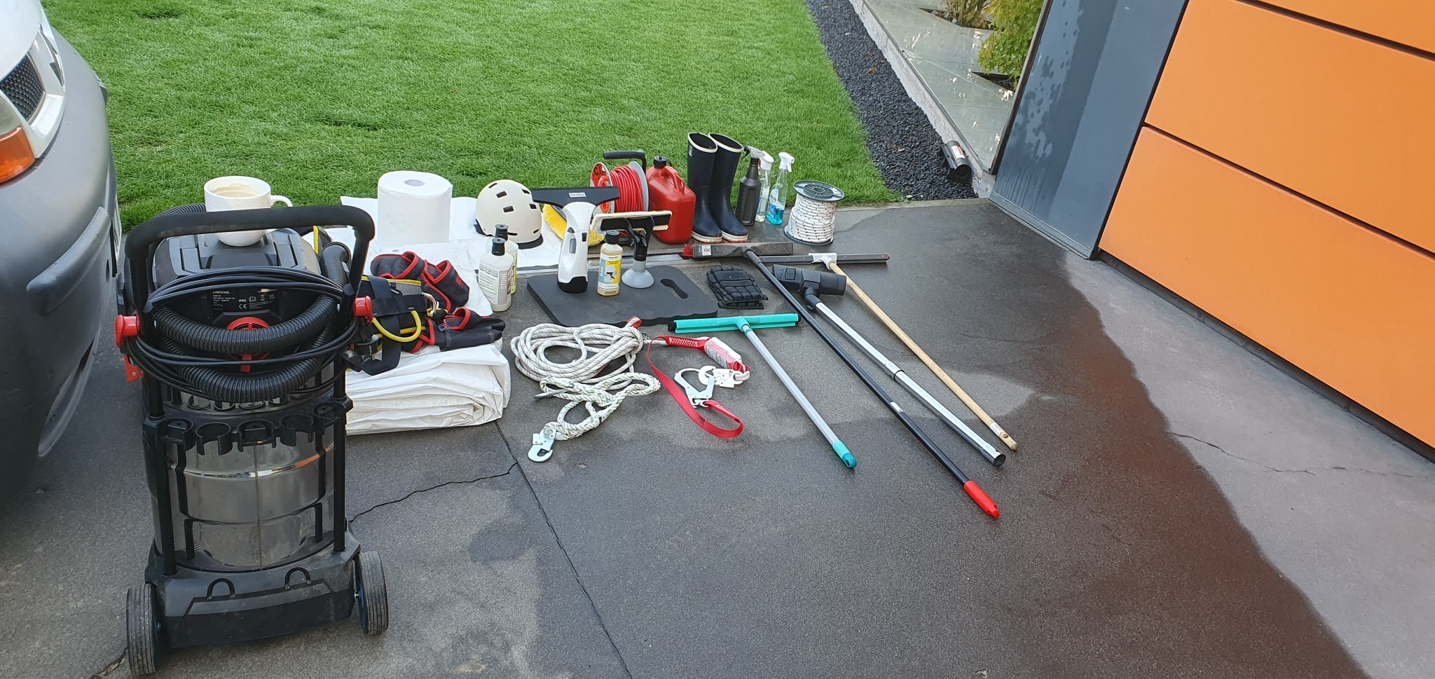 Akcesoria do mycia okien na zewnątrz: odkurzacz do wody, ściągaczki, liny, uprząż, kask, chemia, kalosze i inne narzędzia pracy przy nowoczesnym domu z zielonym trawnikiem.