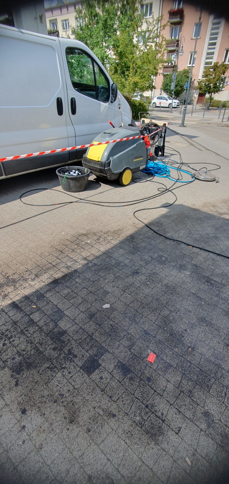 Profesjonalny sprzęt do czyszczenia kostki brukowej Kärcher, z podłączonymi wężami, stoi obok białego busa na chodniku z kostki. W tle budynki miejskie.