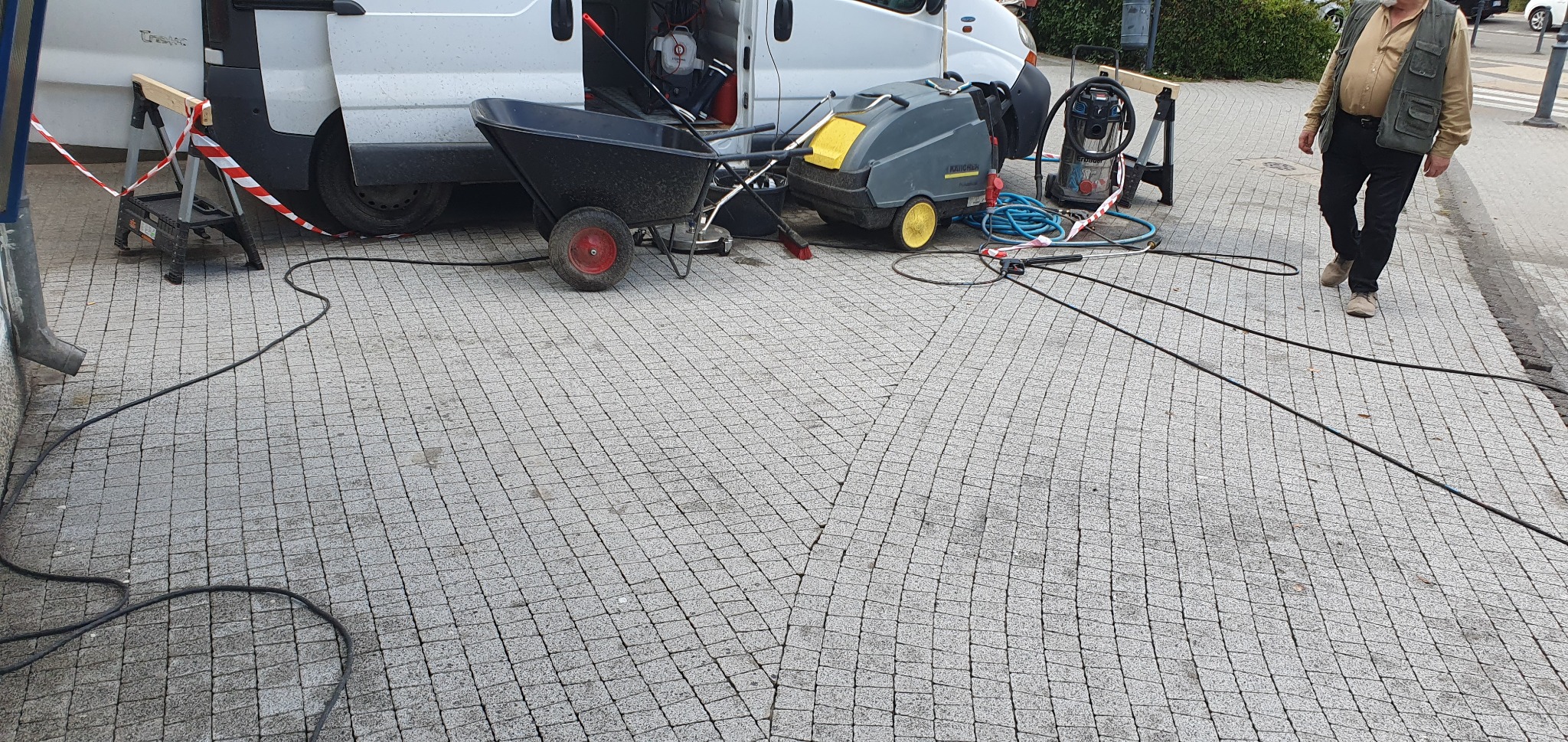 Przygotowanie do czyszczenia kostki brukowej: wózek, myjka ciśnieniowa Karcher i rozłożone węże na szarej kostce. W tle biały bus i osoba w kamizelce.