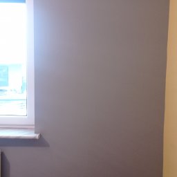 Firma Budowlana Krzysztof Markiewicz - Świeżo pomalowana szara ściana w pokoju z drewnianą podłogą, widoczny fragment okna z białymi ramami i karnisz.
