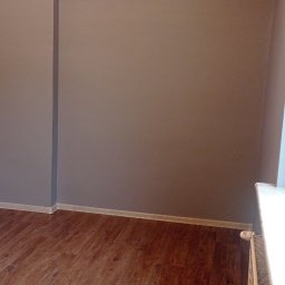 Firma Budowlana Krzysztof Markiewicz - Wnętrze pokoju po remoncie: szare ściany, drewniana podłoga, białe listwy przypodłogowe i grzejnik. Widoczny kratka wentylacyjna na ścianie.