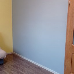 Firma Budowlana Krzysztof Markiewicz - Pomieszczenie po odświeżeniu: ściana pomalowana na szaro, kontrastująca z żółtą ścianą boczną, widoczna drewniana stolarka drzwiowa i panelowa podłoga.