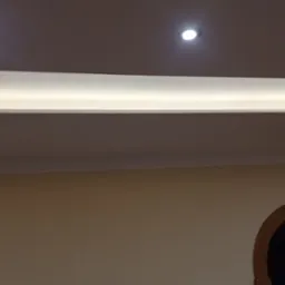 Wykończony sufit podwieszany z oświetleniem LED, widoczne halogeny i dekoracyjny element wpuszczony w sufit. Ściany pomalowane na jasny kolor, widoczny fragment łuku drzwiowego.