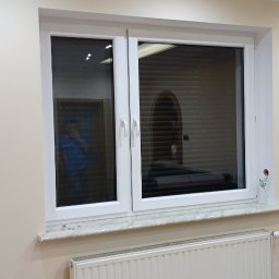 Firma Budowlana Krzysztof Markiewicz - Wnętrze pokoju po remoncie: białe okno PCV z marmurowym parapetem nad grzejnikiem panelowym, ściany pomalowane na jasny kremowy odcień, podłoga wyłożona panelami imitującymi drewno.