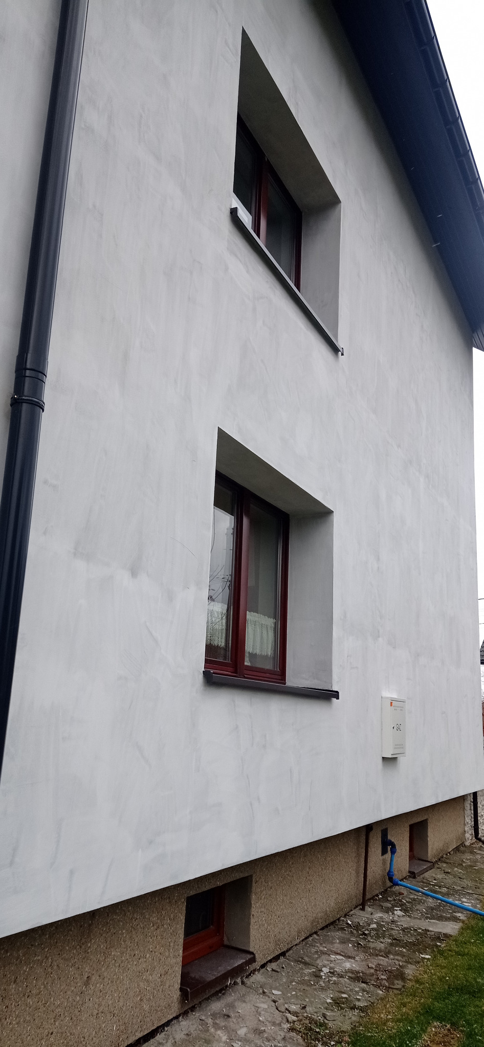 Świeżo otynkowana elewacja domu jednorodzinnego z widocznymi oknami w brązowych ramach, rurą spustową i skrzynką gazową. Na dole widoczny fragment izolacji fundamentów.