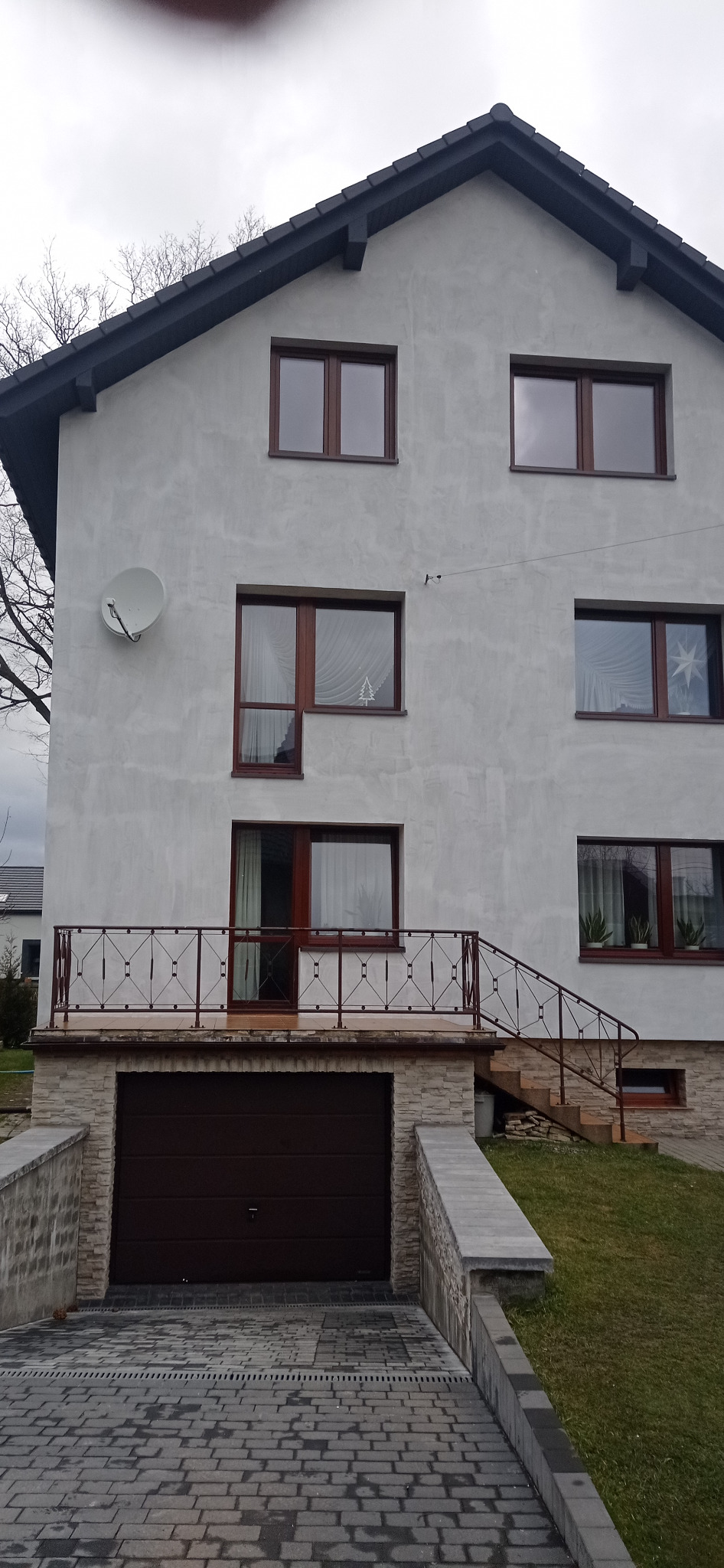 Dwukondygnacyjny dom jednorodzinny z szarą elewacją, brązowymi oknami i garażem wbudowanym w bryłę budynku, widoczny z poziomu podjazdu z kostki brukowej.