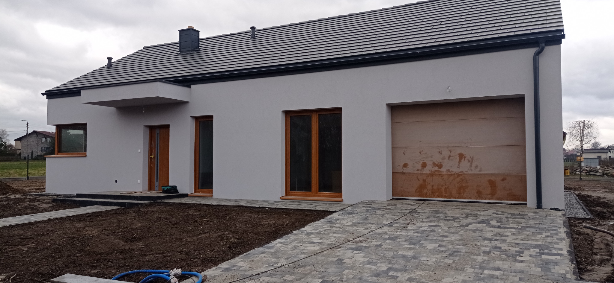 Nowoczesny dom jednorodzinny z szarą elewacją, brązowymi oknami i bramą garażową, widoczny chodnik z kostki brukowej i przygotowany teren pod ogród, pochmurne niebo.