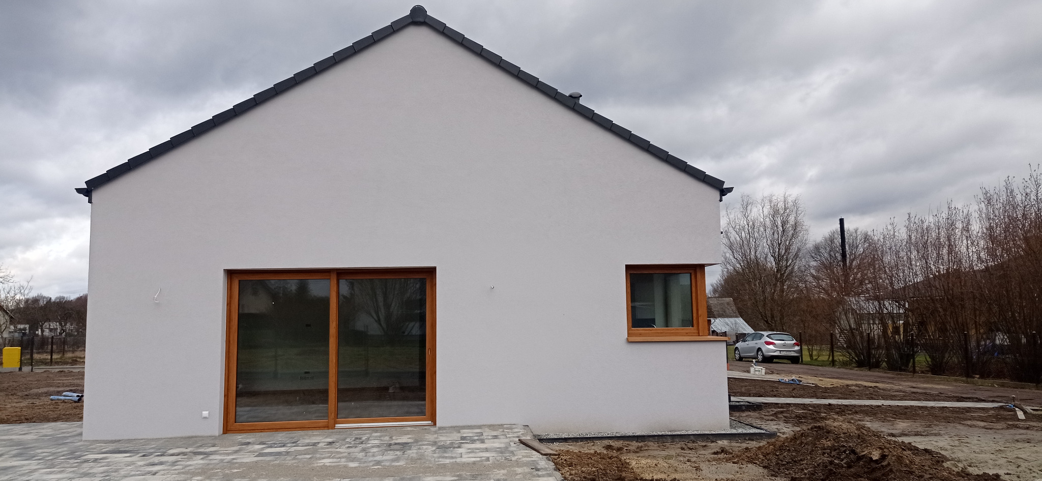 Wykończona elewacja domu jednorodzinnego w kolorze jasnoszarym, z widocznymi drewnianymi oknami i drzwiami przesuwnymi, oraz fragmentem ułożonej kostki brukowej na podwórku. W tle widoczny samochód...
