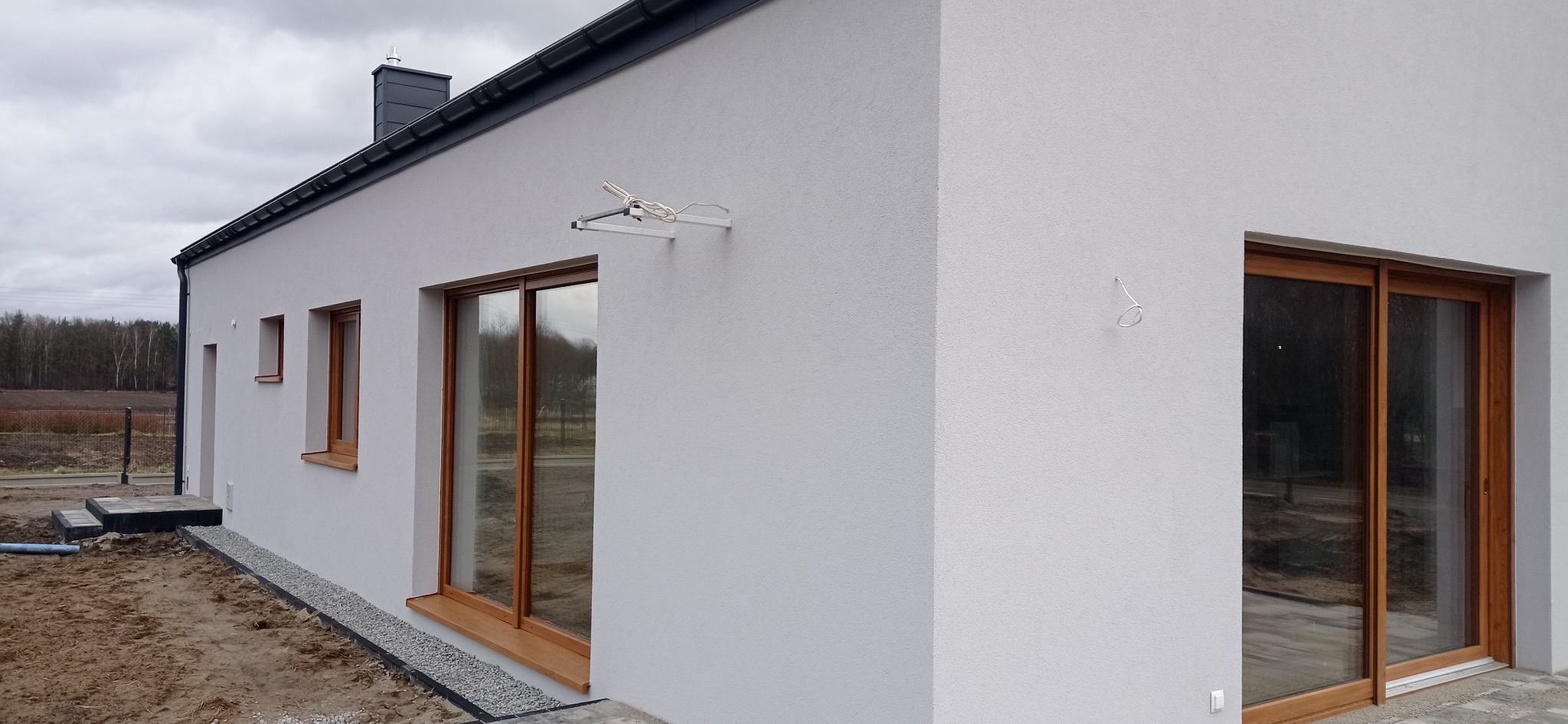 Fragment elewacji domu jednorodzinnego z drewnianą stolarką okienną, widoczna antena satelitarna na ścianie i fragment terenu wokół budynku.