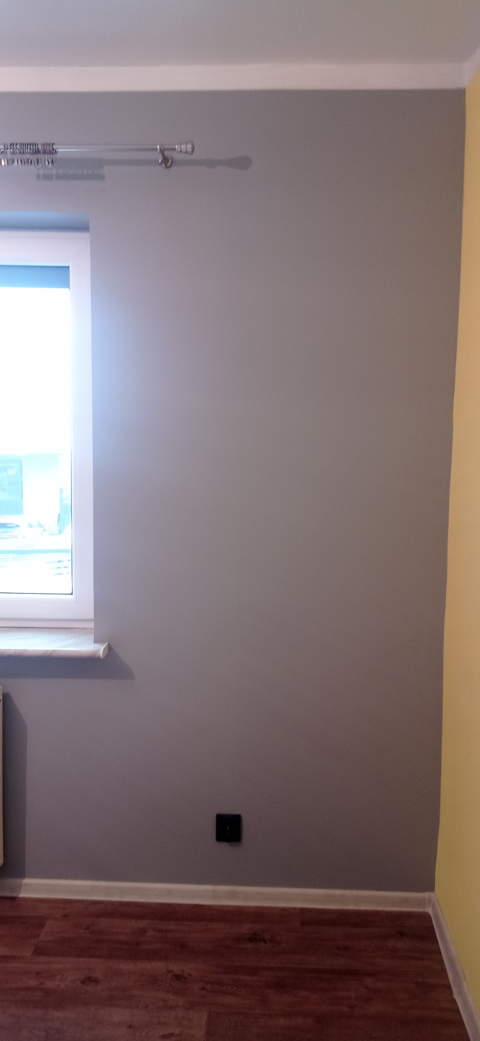 Świeżo pomalowana szara ściana w pokoju z drewnianą podłogą, widoczny fragment okna z białymi ramami i karnisz.