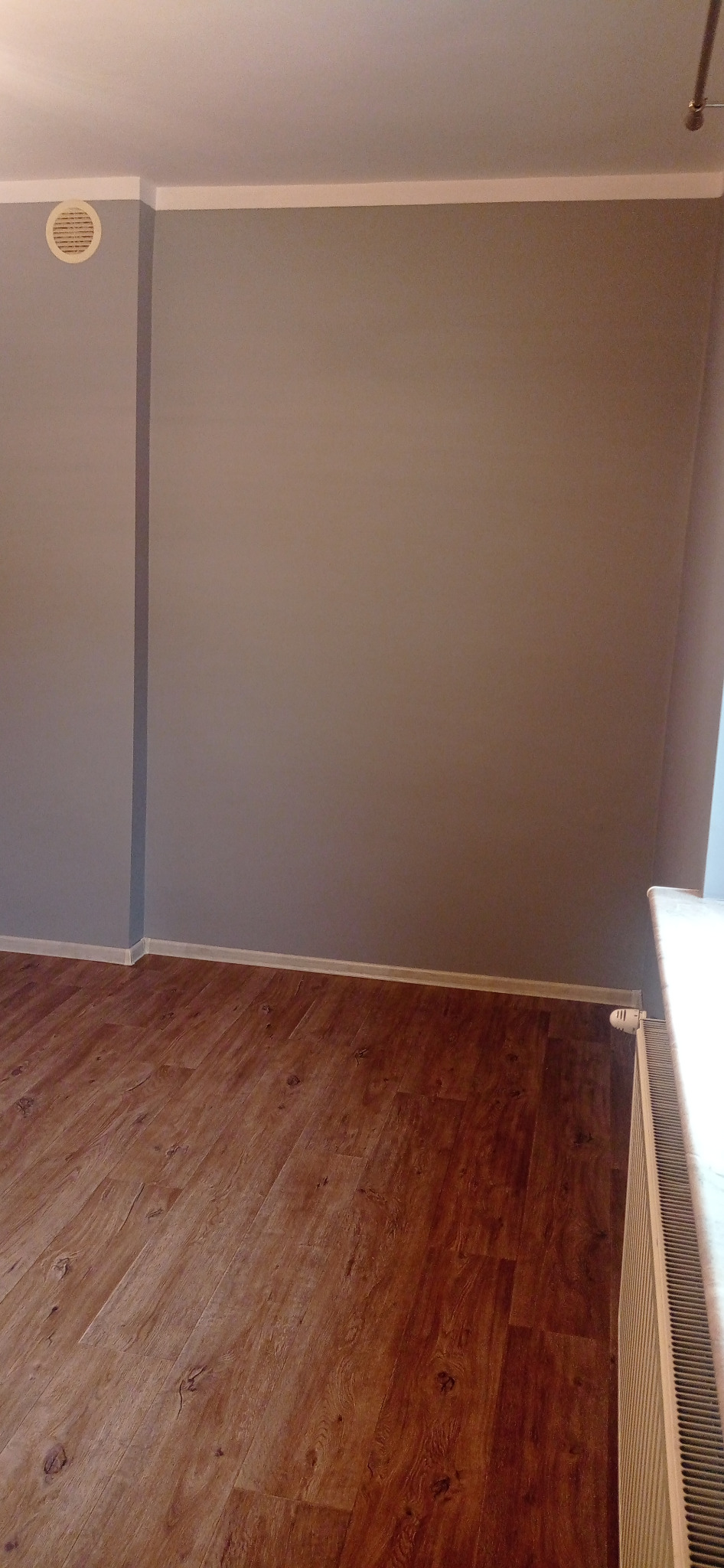 Wnętrze pokoju po remoncie: szare ściany, drewniana podłoga, białe listwy przypodłogowe i grzejnik. Widoczny kratka wentylacyjna na ścianie.