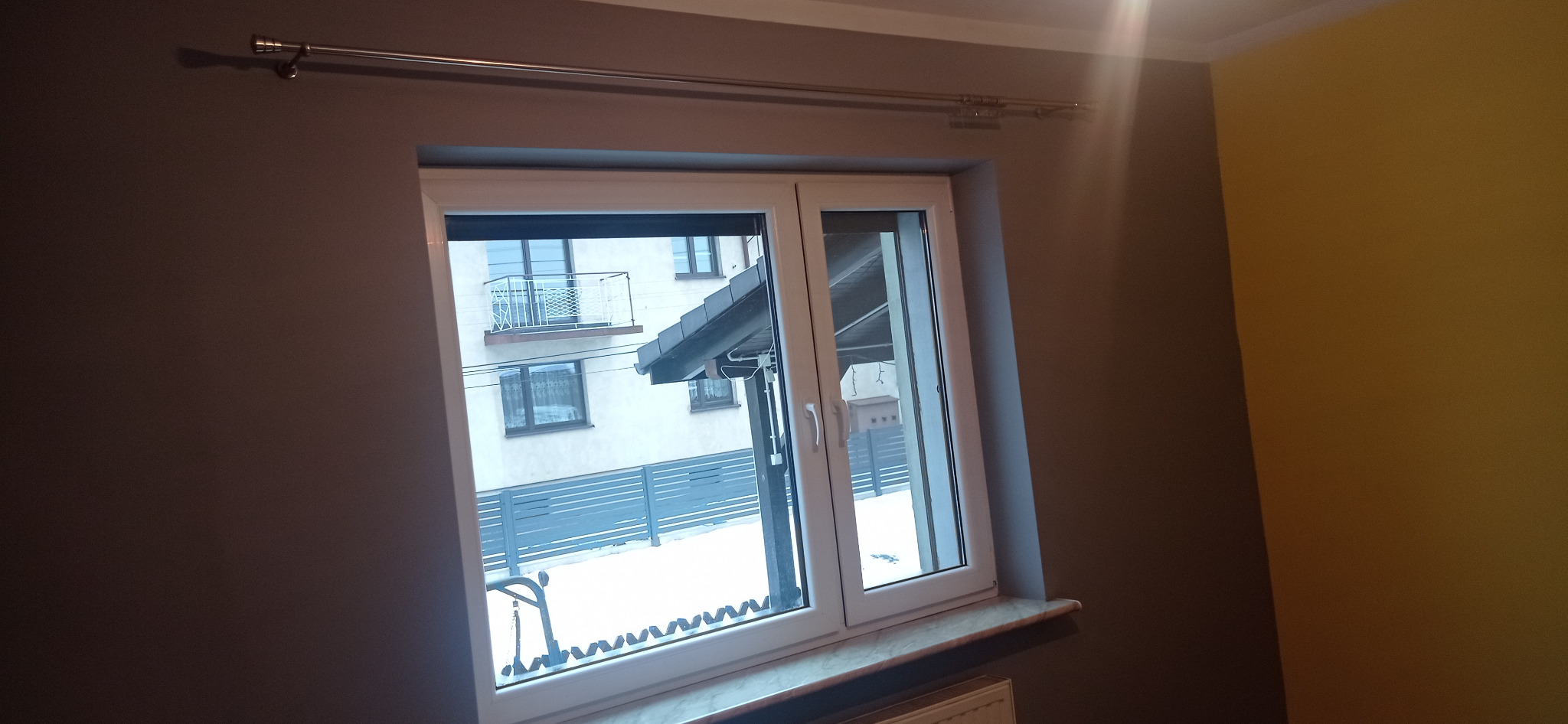 Świeżo pomalowane, gładkie ściany w odcieniach szarości i żółci, widoczne w kadrze z oknem PCV z roletą, przez które widać ośnieżony krajobraz i budynek wielorodzinny z balkonami.