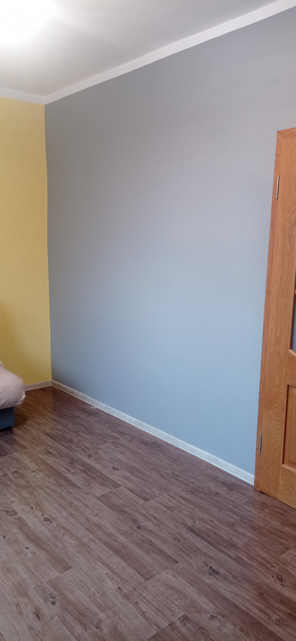 Pomieszczenie po odświeżeniu: ściana pomalowana na szaro, kontrastująca z żółtą ścianą boczną, widoczna drewniana stolarka drzwiowa i panelowa podłoga.