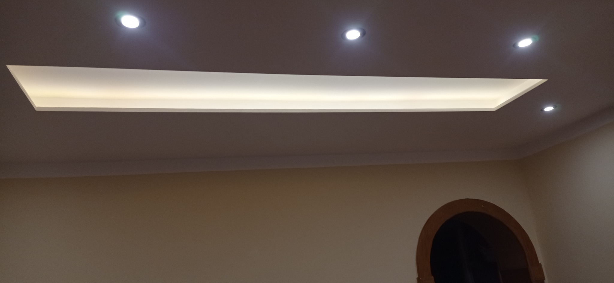 Wykończony sufit podwieszany z oświetleniem LED, widoczne halogeny i dekoracyjny element wpuszczony w sufit. Ściany pomalowane na jasny kolor, widoczny fragment łuku drzwiowego.