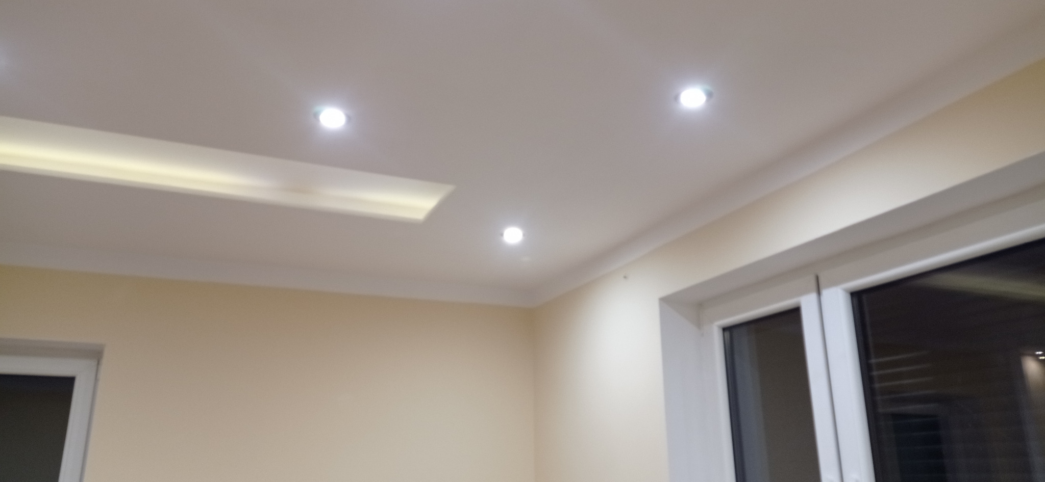 Pomieszczenie po malowaniu ścian na jasny kremowy kolor, z zamontowanym oświetleniem sufitowym punktowym i dekoracyjnym podwieszanym sufitem z oświetleniem LED, widoczne okno z białymi ramami.