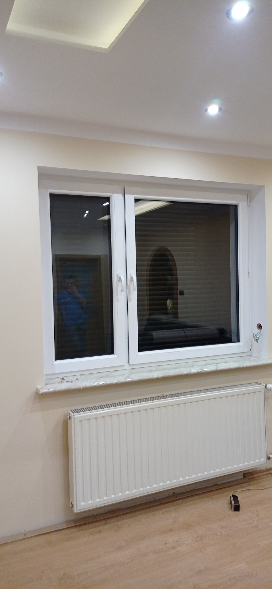 Wnętrze pokoju po remoncie: białe okno PCV z marmurowym parapetem nad grzejnikiem panelowym, ściany pomalowane na jasny kremowy odcień, podłoga wyłożona panelami imitującymi drewno.