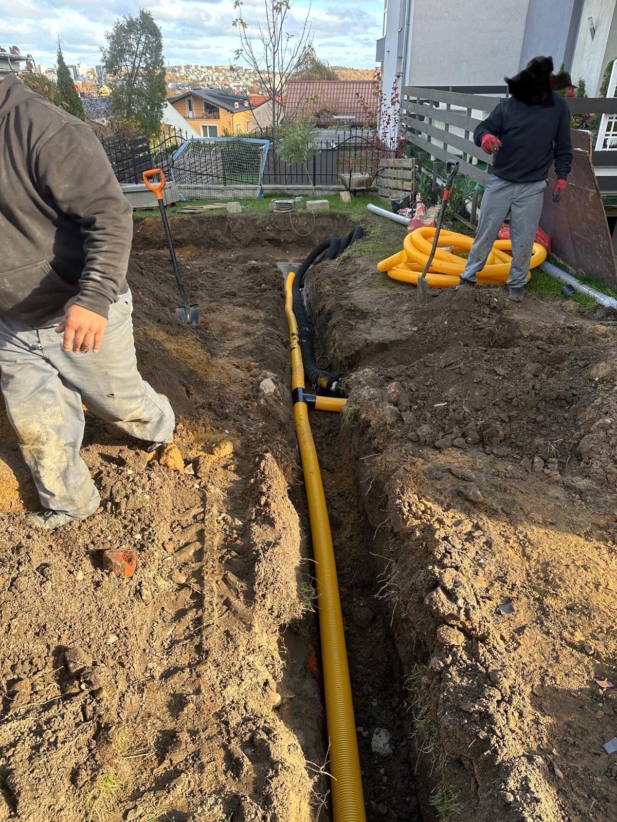 Wykop w ziemi z ułożonymi żółtymi rurami drenażowymi, widoczne ślady opon, dwóch mężczyzn w roboczych ubraniach i łopaty w tle, domy w oddali.