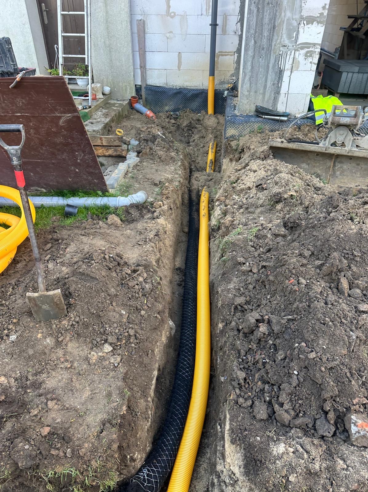 Wykop w ziemi z ułożonymi rurami drenażowymi w kolorze żółtym i czarnym, widoczne narzędzia i fragment budynku w tle.