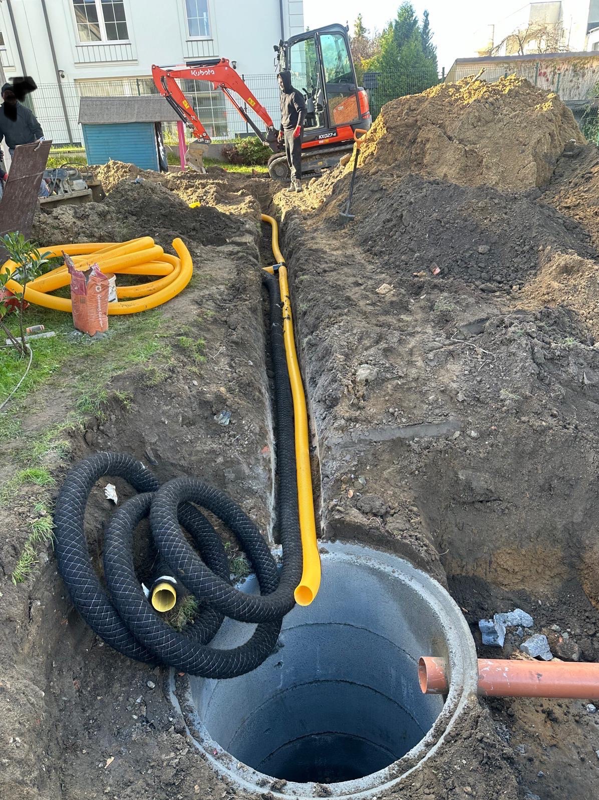 Głęboki wykop z rurami drenażowymi w ziemi, w tle minikoparka Kubota i pracownicy, widoczny betonowy krąg studzienny z podłączonymi rurami.