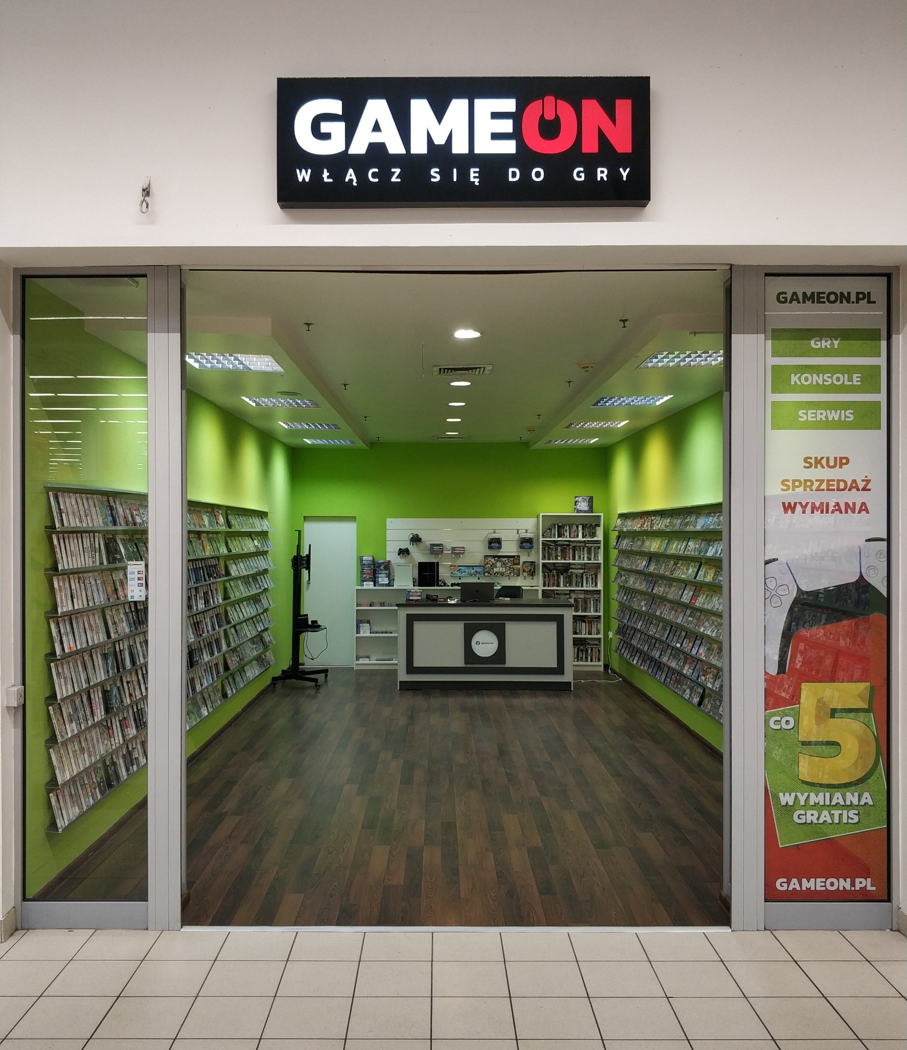 Wejście do sklepu z grami GAMEON, półki wypełnione grami po bokach, jasne wnętrze z zielonymi ścianami i drewnianą podłogą, plakat z ofertą wymiany gier na szybie.