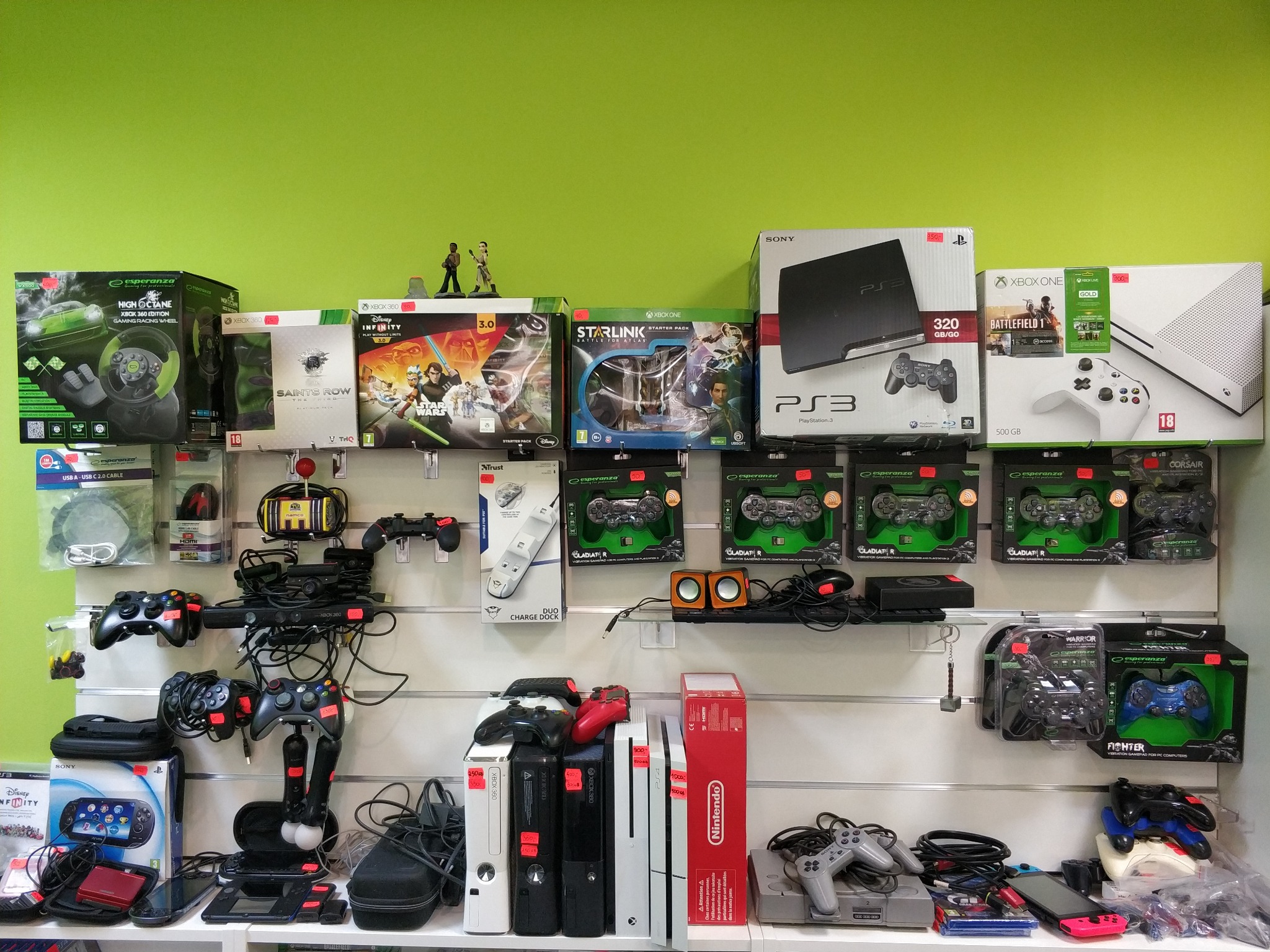 Półka w sklepie z elektroniką, prezentująca konsole Xbox, PlayStation, Nintendo, gry, kontrolery oraz akcesoria gamingowe na tle zielonej ściany. Widoczne modele konsol PS3, Xbox 360, Xbox One...