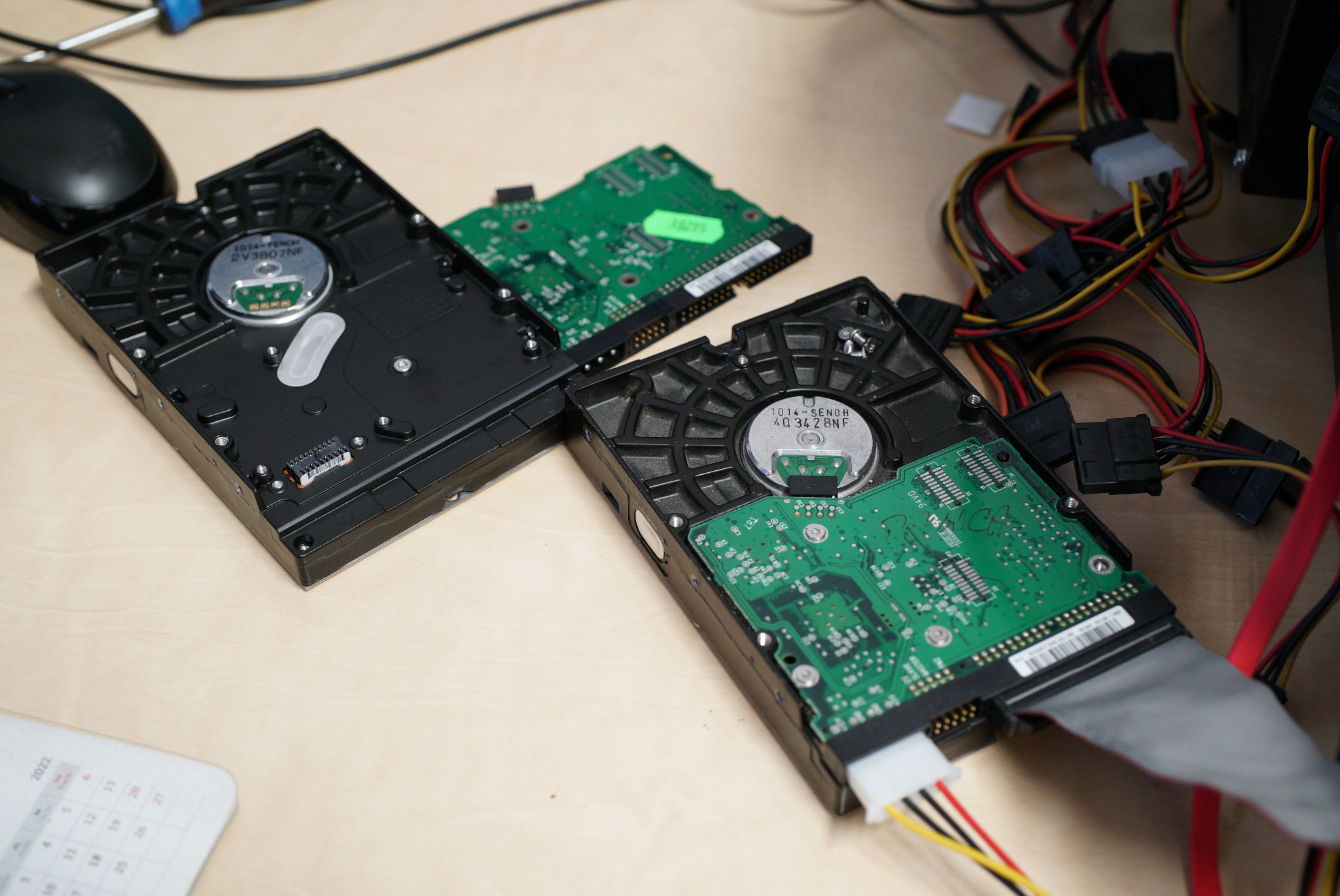 Otwarty dysk twardy z widoczną elektroniką, kable zasilające i data kabel na drewnianym biurku, obok leży kalendarz.