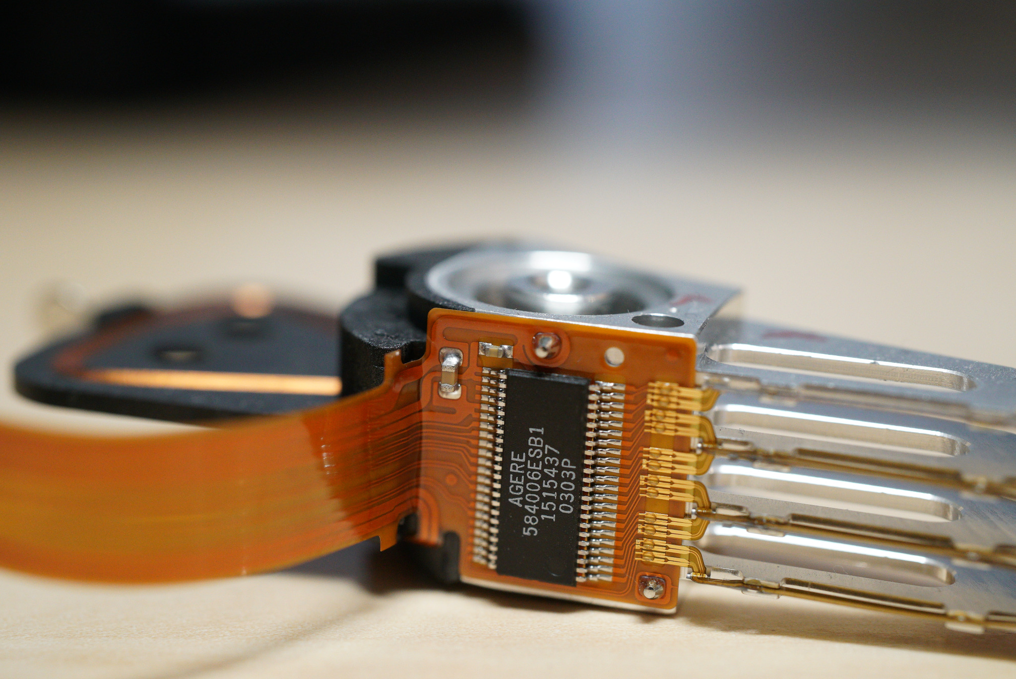 Głowica dysku twardego z widocznym układem scalonym AGERE 584006ESB1 1515437 0303P, podłączona za pomocą pomarańczowej taśmy, leżąca na jasnym blacie.