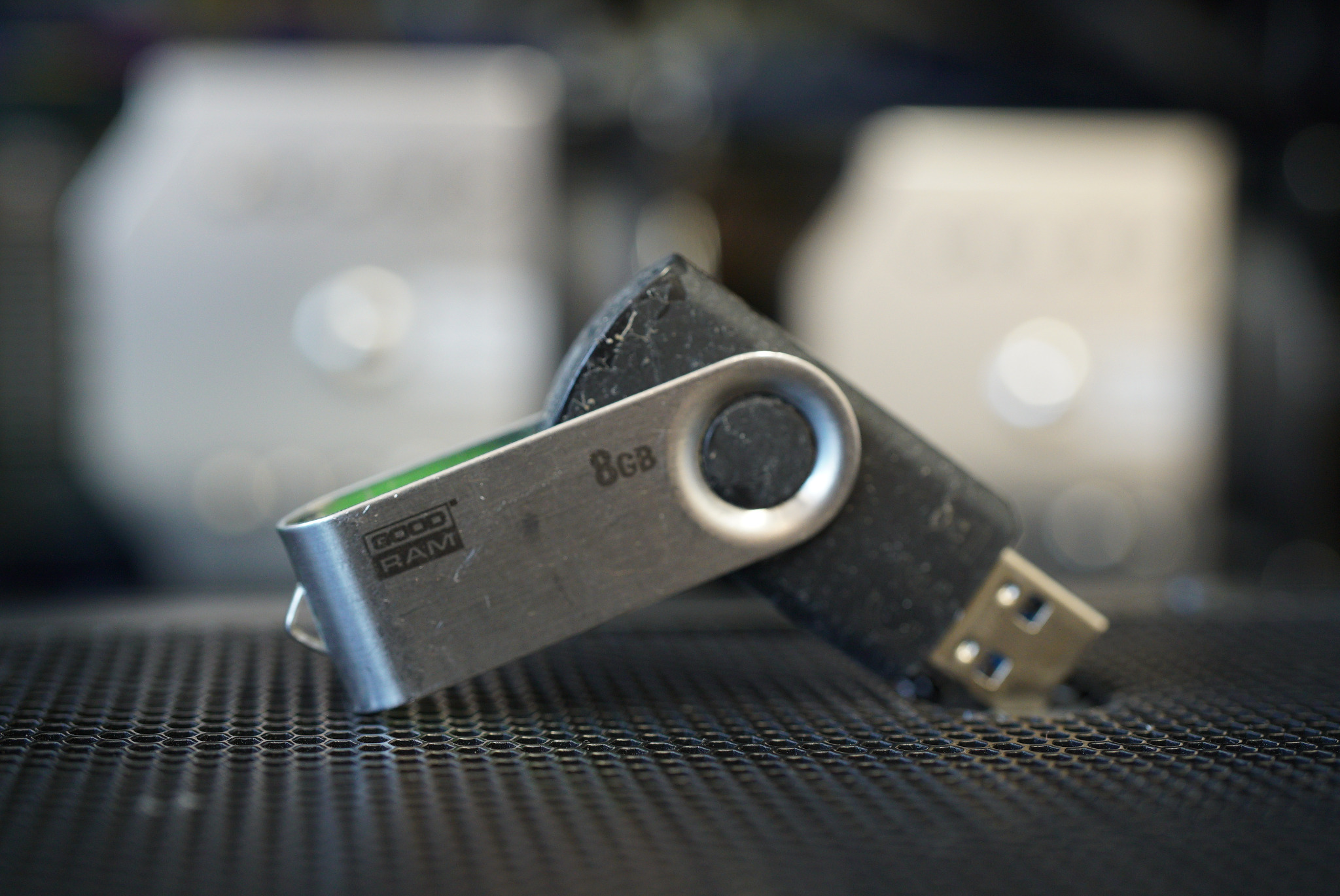 Czarny pendrive 8GB z metalową obudową leżący na perforowanej powierzchni, z widocznymi śladami użytkowania i logiem 'GOOD RAM'. W tle rozmyte obudowy komputerowe.