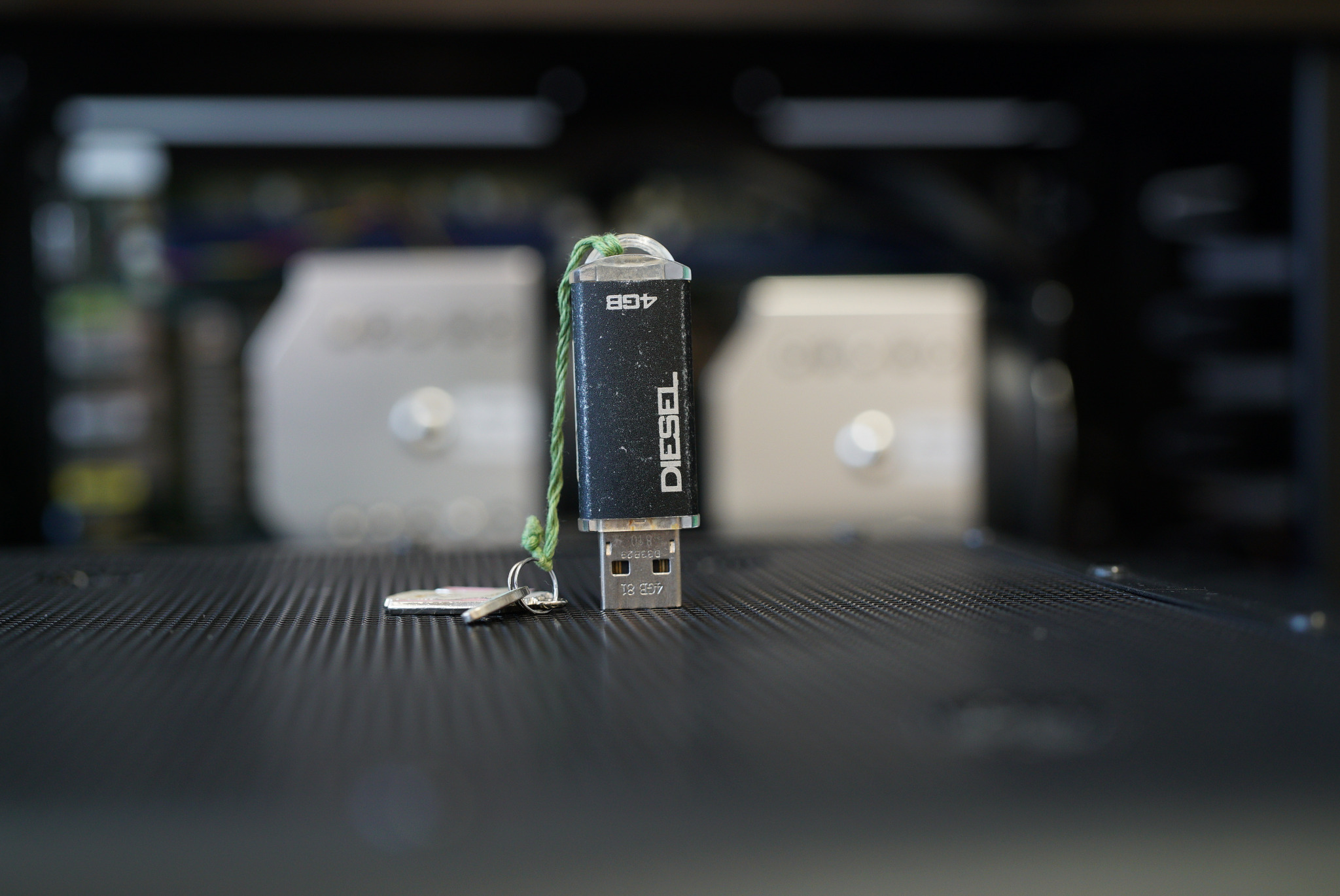 Czarny pendrive marki Diesel o pojemności 4GB, z zielonym sznurkiem, leży na czarnej, perforowanej powierzchni wewnątrz obudowy komputera, obok kluczyk do pendrive.