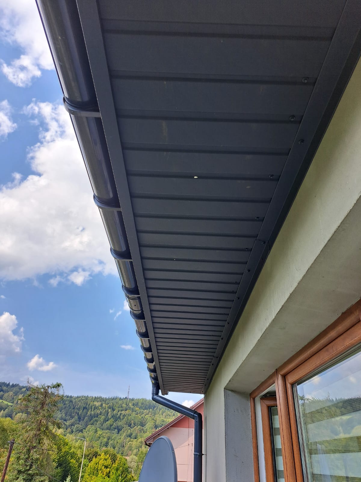 Ciemnoszary dach z blachy trapezowej i system rynnowy w kolorze, widok z dołu na tle błękitnego nieba z chmurami i zielonych wzgórz. Nowoczesne wykończenie.