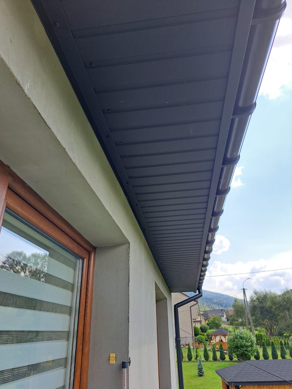 Grafitowa podbitka dachowa z rynnami, widok z dołu, z fragmentem okna i elewacji. W tle zieleń i zabudowa jednorodzinna.