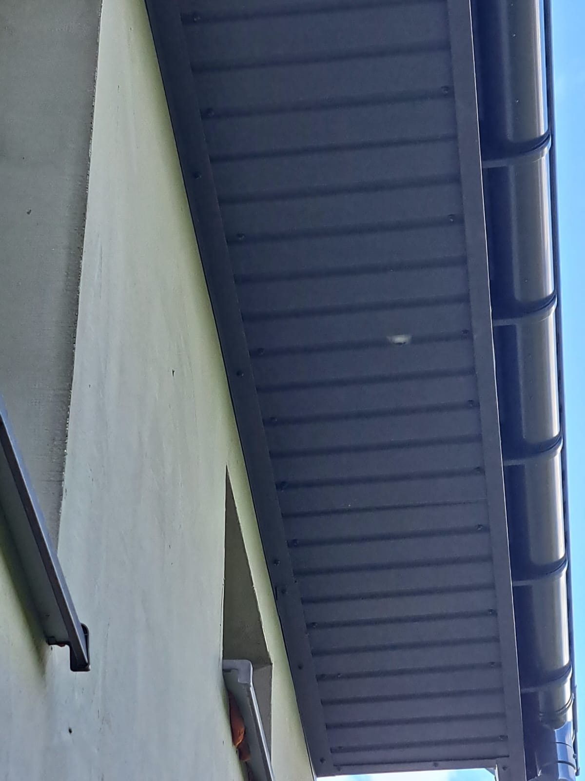Ciemna podbitka dachowa z widoczną rynną, zamontowana przy ścianie budynku. Ujęcie z dołu, fragment błękitnego nieba w tle. Widoczny fragment ściany budynku.