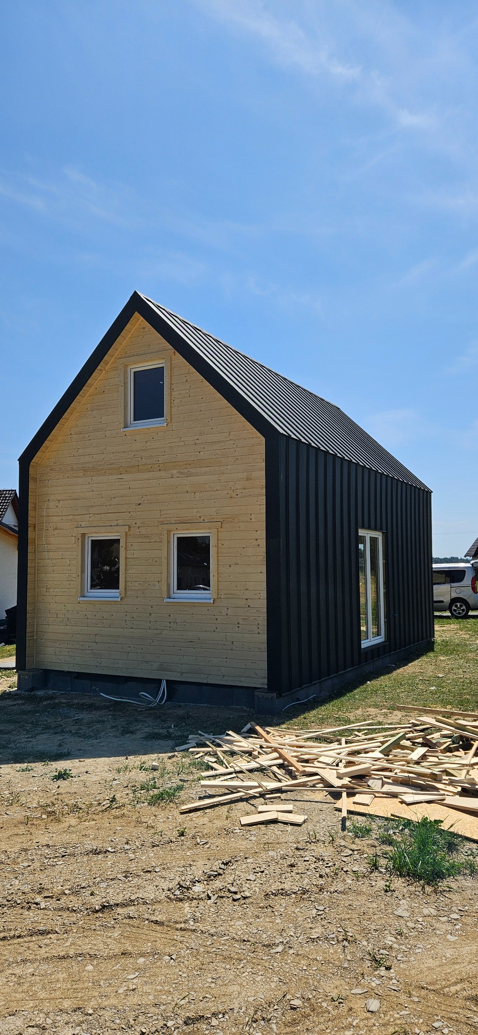 Nowoczesny dom z dachem dwuspadowym, wykończony drewnem i czarną blachą trapezową. Widoczne okna i fragment terenu z resztkami drewna po budowie.